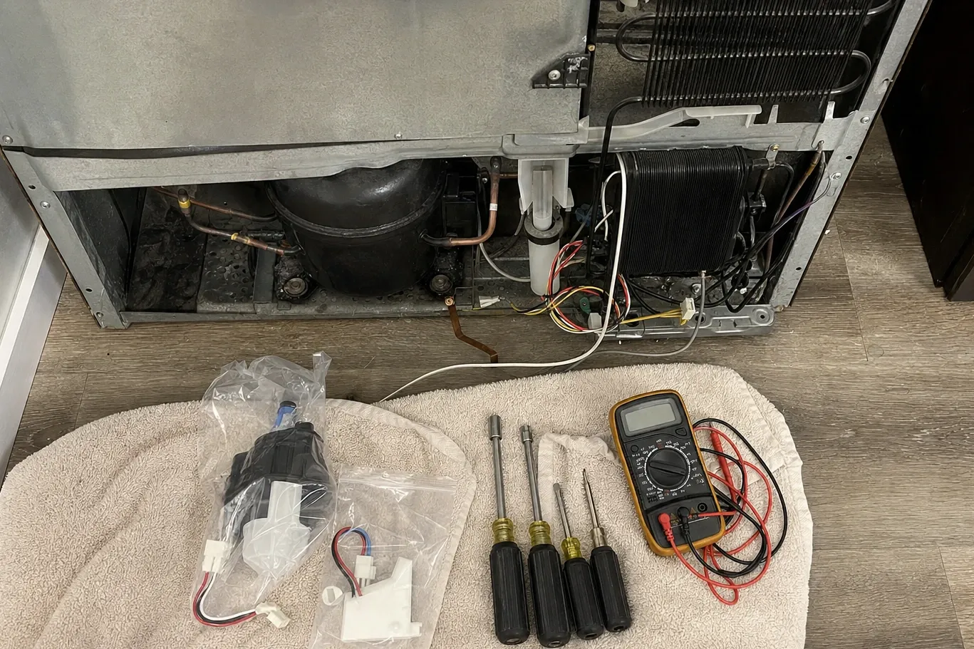 Bagged replacement parts, nut drivers and a multimeter laid out on a towel behind a unit pulled from the wall, compressor and condenser exposed